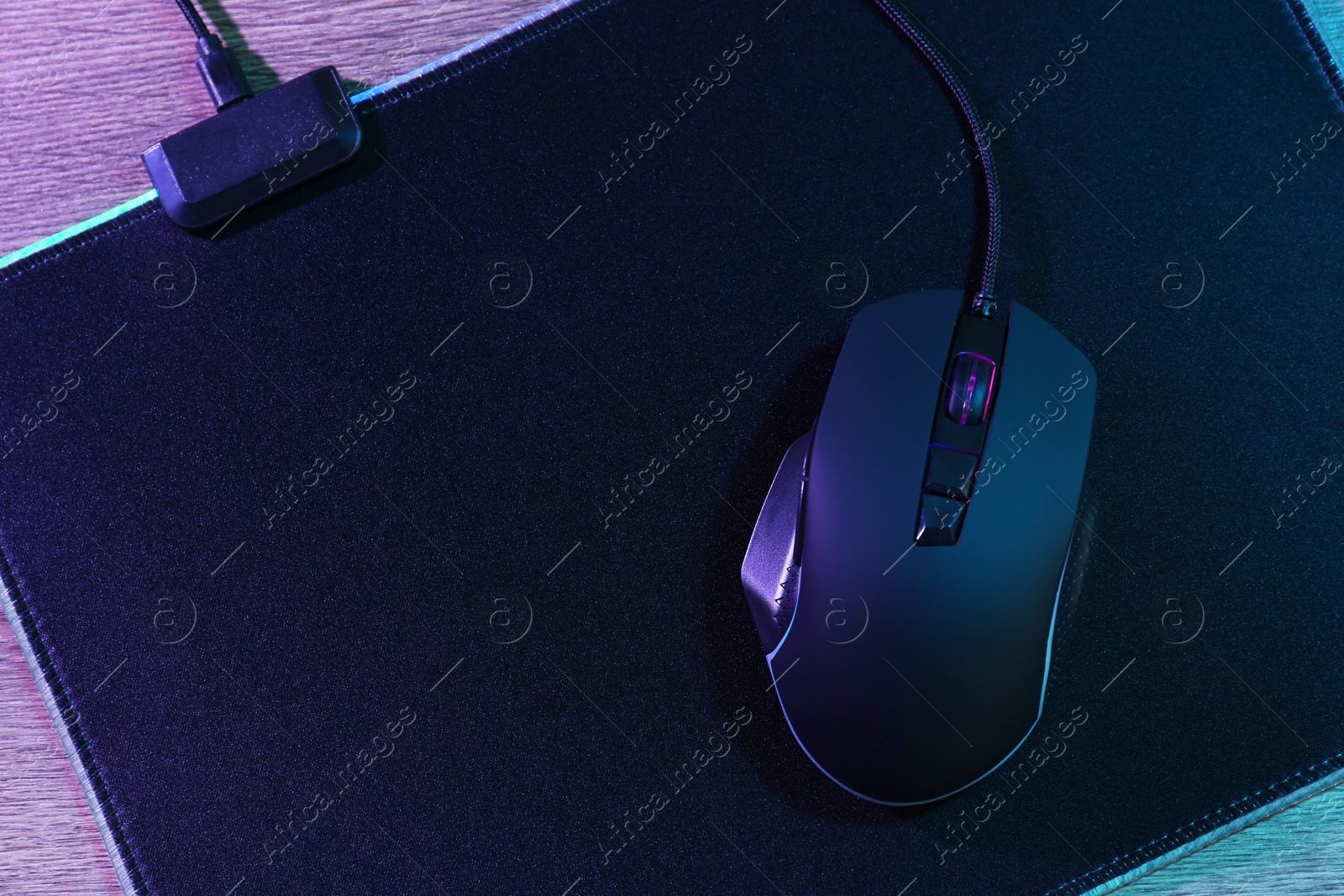 Photo of Black computer mouse and mousepad on wooden table in neon lights, top view