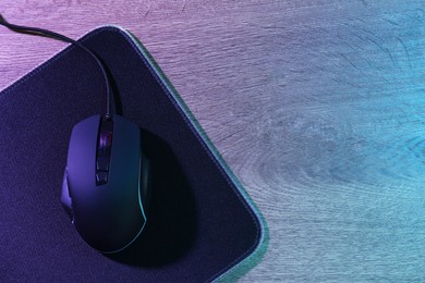 Photo of Black computer mouse and mousepad on wooden table in neon lights, top view. Space for text