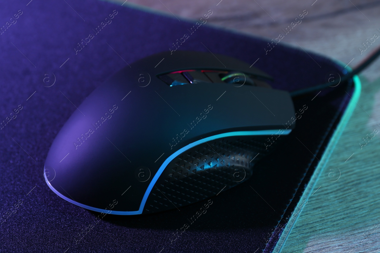 Photo of Black computer mouse and mousepad on wooden table in neon lights, closeup