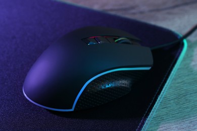 Photo of Black computer mouse and mousepad on wooden table in neon lights, closeup