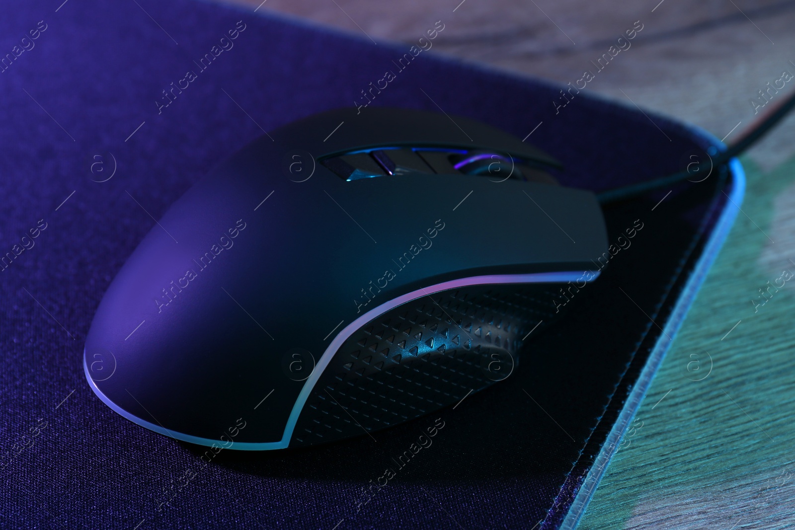 Photo of Black computer mouse and mousepad on wooden table in neon lights, closeup