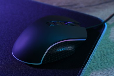 Photo of Black computer mouse and mousepad on wooden table in neon lights, closeup