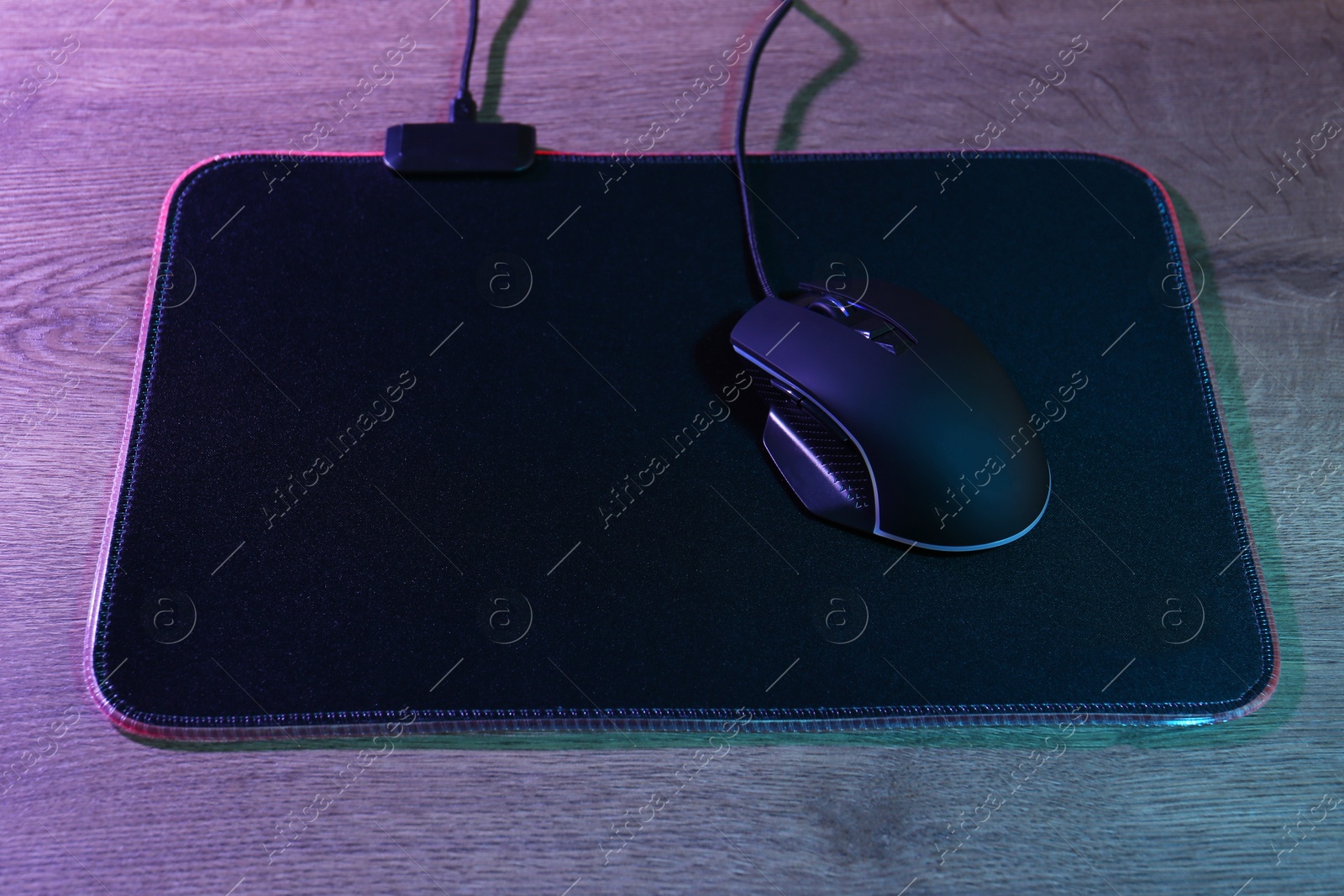 Photo of Black computer mouse and mousepad on wooden table in neon lights