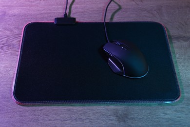 Photo of Black computer mouse and mousepad on wooden table in neon lights