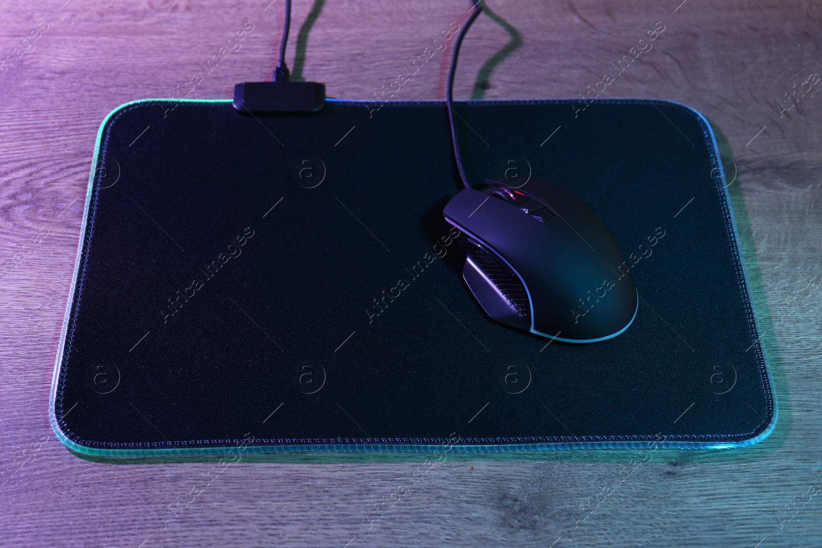 Photo of Black computer mouse and mousepad on wooden table in neon lights