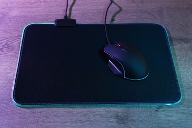 Photo of Black computer mouse and mousepad on wooden table in neon lights