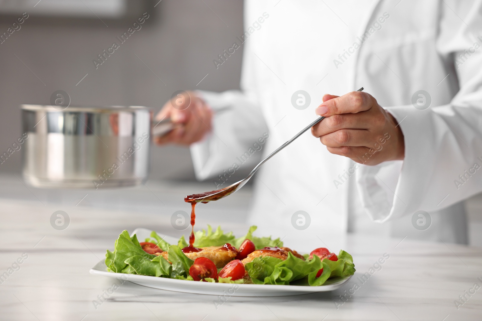 Photo of Professional chef adding sauce to dish with baked chicken at white marble table in kitchen, closeup