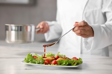 Professional chef adding sauce to dish with baked chicken at white marble table in kitchen, closeup