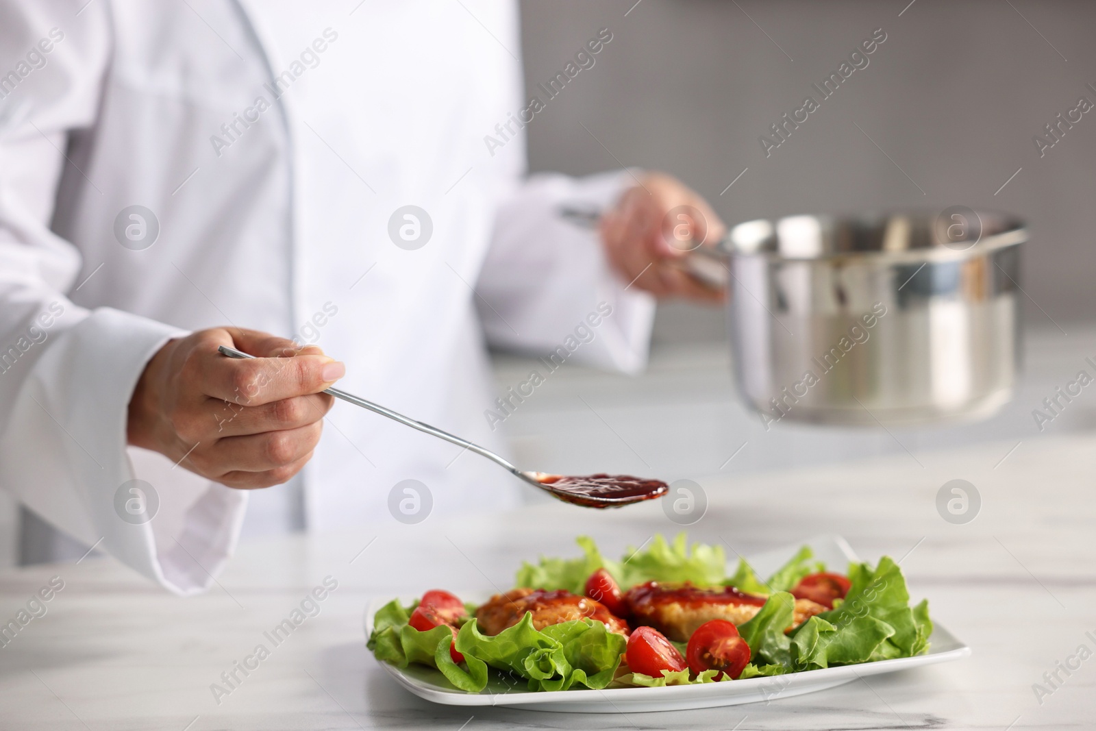 Photo of Professional chef adding sauce to dish with baked chicken at white marble table in kitchen, closeup