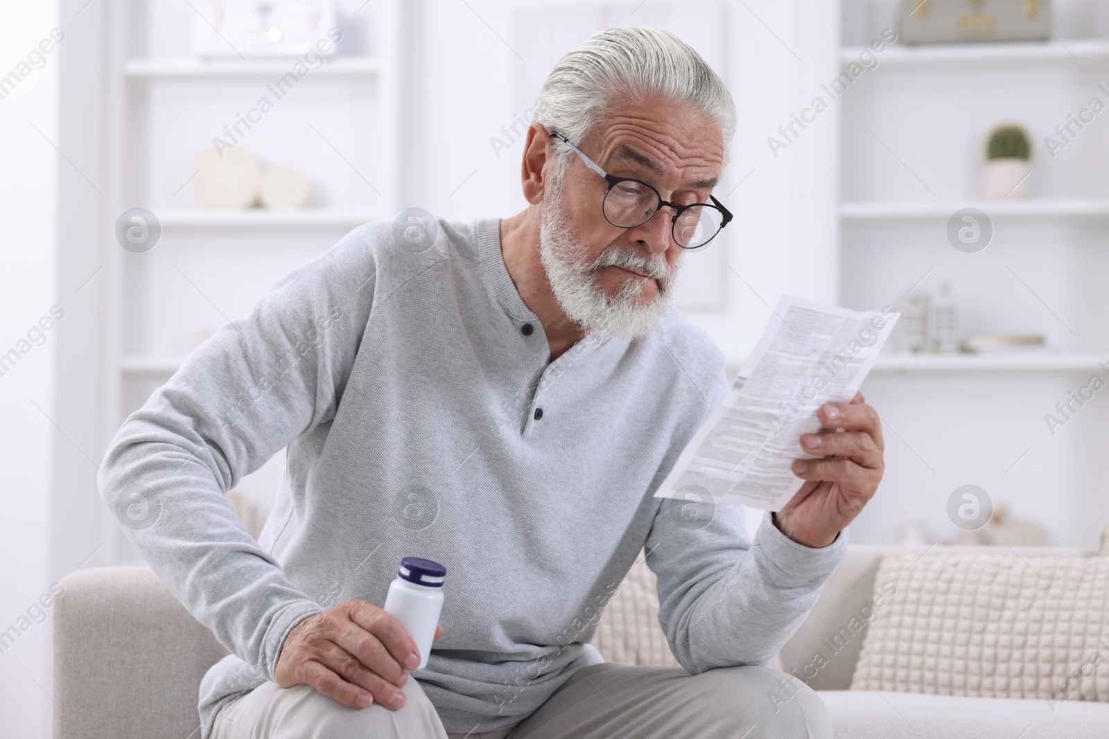 Photo of Senior man with pills reading medicine instruction on sofa at home