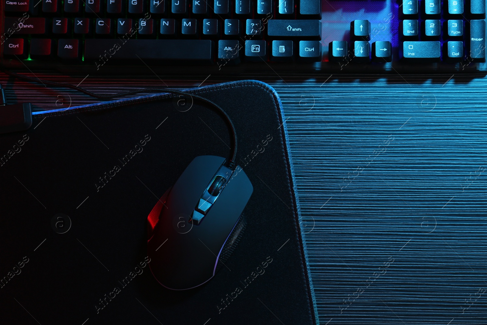 Photo of Computer mouse, mousepad and keyboard on dark wooden table in neon lights, flat lay. Space for text