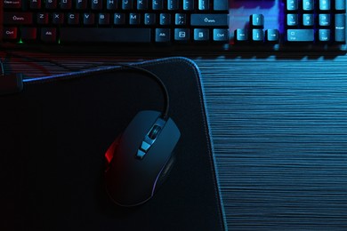 Photo of Computer mouse, mousepad and keyboard on dark wooden table in neon lights, flat lay. Space for text