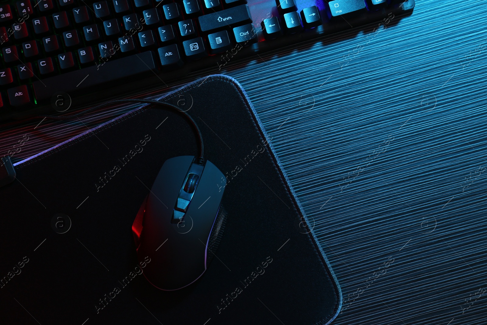 Photo of Computer mouse, mousepad and keyboard on dark wooden table in neon lights, flat lay. Space for text