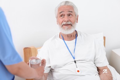 Photo of Senior man with emergency call button taking glass of water nurse giving him in hospital, closeup