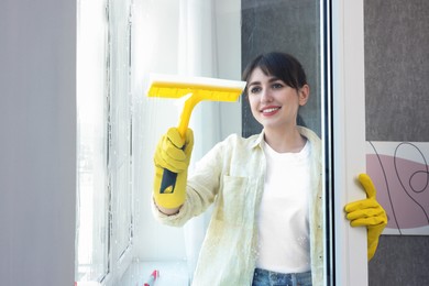 Beautiful young woman with squeegee cleaning window indoors