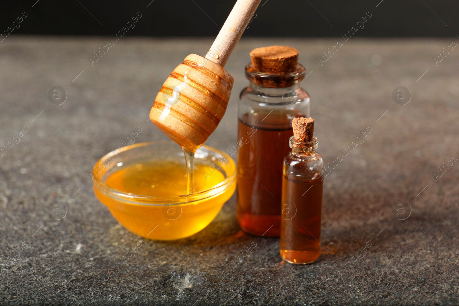 Photo of Pouring honey from dipper into bowl and bottles of tincture on grey textured table, closeup. Alternative medicine