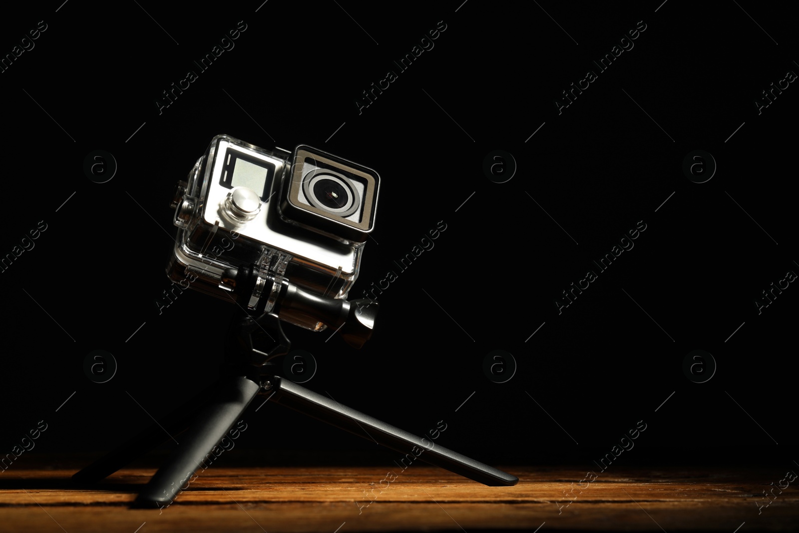 Photo of Modern action camera and tripod on wooden surface against black background. Space for text