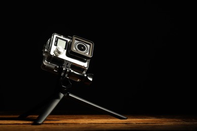 Modern action camera and tripod on wooden surface against black background. Space for text