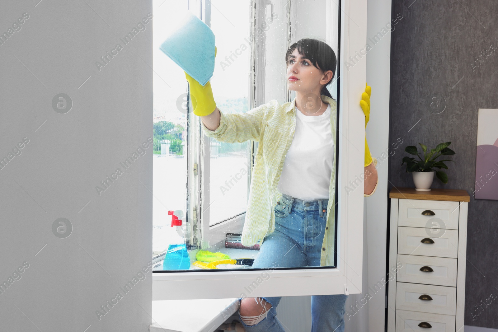 Photo of Beautiful young woman with napkin cleaning window indoors