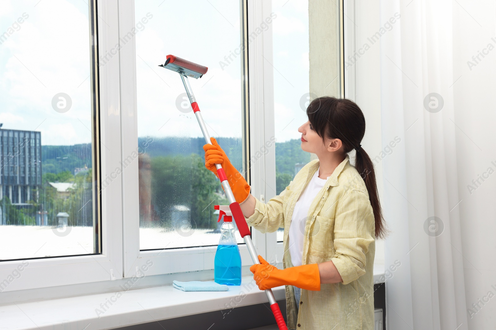 Photo of Beautiful young woman with squeegee tool cleaning window indoors