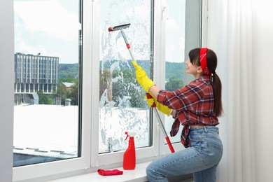 Young woman with squeegee tool cleaning window indoors