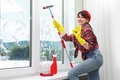 Beautiful young woman with squeegee tool showing OK gesture while cleaning window indoors