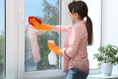 Woman with spray bottle of detergent and napkin cleaning window indoors