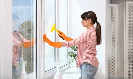 Beautiful young housewife with squeegee cleaning window indoors