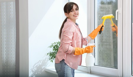 Photo of Beautiful young woman with squeegee and spray bottle of detergent cleaning window indoors