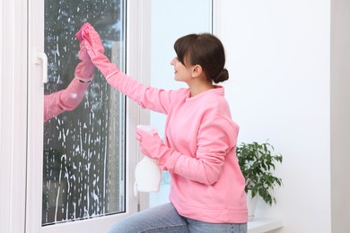 Beautiful young woman with spray bottle of detergent and napkin cleaning window indoors