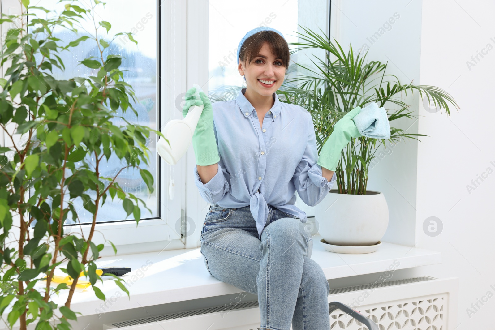 Photo of Cleaning window. Beautiful young woman with spray bottle of detergent and napkin on windowsill indoors