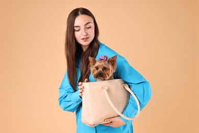 Beautiful young woman carrying cute Yorkshire Terrier dog in bag on beige background