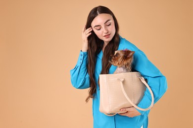 Beautiful young woman carrying cute Yorkshire Terrier dog in bag on beige background