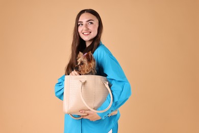 Photo of Beautiful young woman carrying cute Yorkshire Terrier dog in bag on beige background