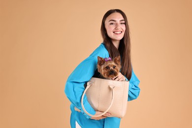 Beautiful young woman carrying cute Yorkshire Terrier dog in bag on beige background