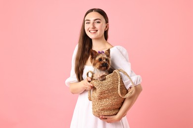 Beautiful young woman holding wicker bag with cute Yorkshire Terrier dog on pink background