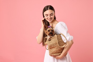 Beautiful young woman holding wicker bag with cute Yorkshire Terrier dog on pink background