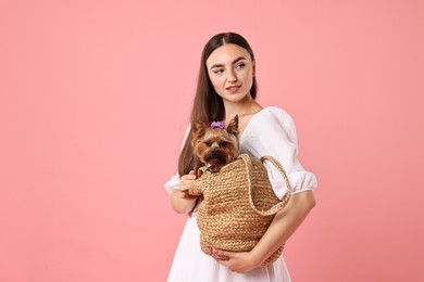 Beautiful young woman holding wicker bag with cute Yorkshire Terrier dog on pink background