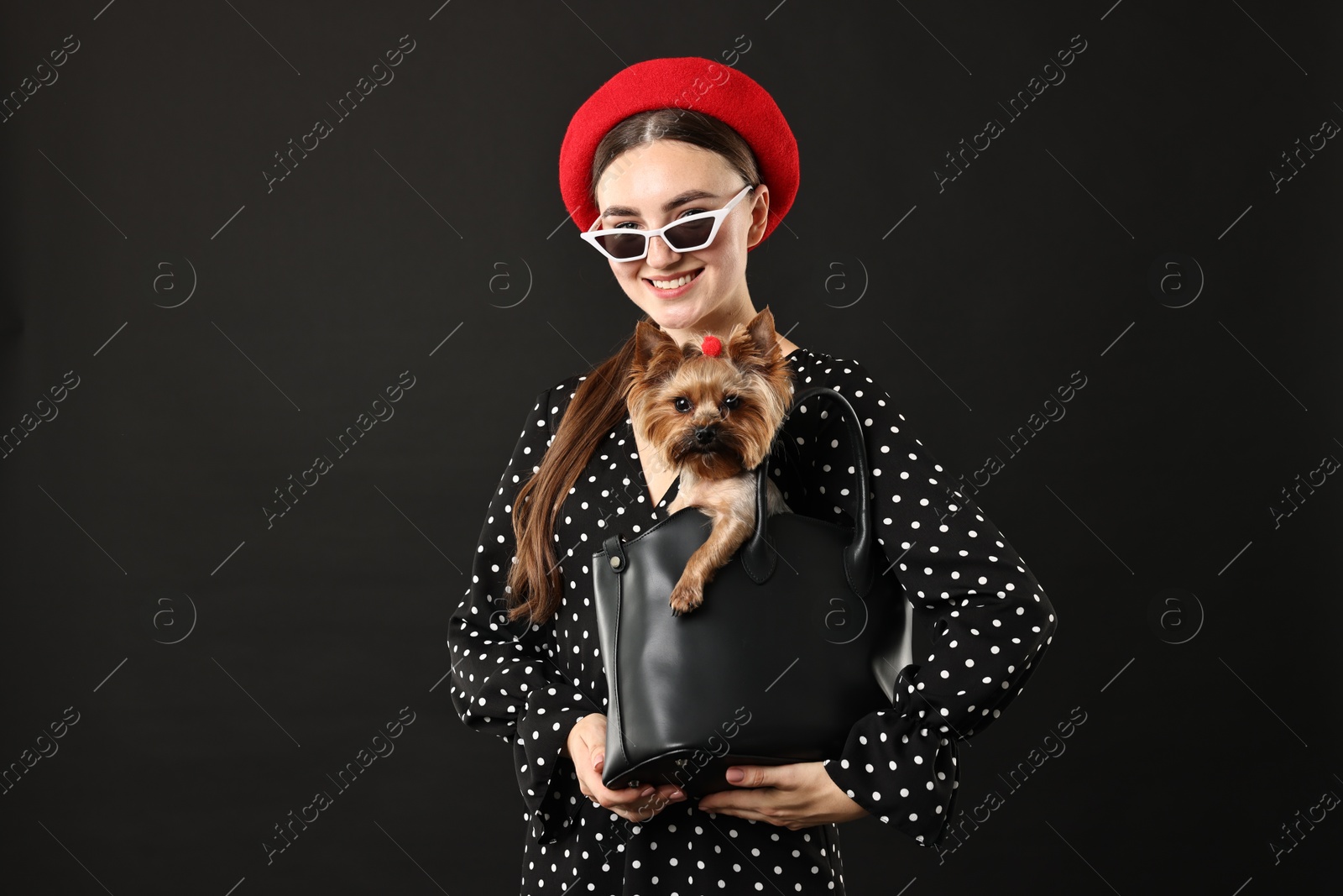 Photo of Woman in sunglasses holding bag with cute Yorkshire Terrier dog on black background