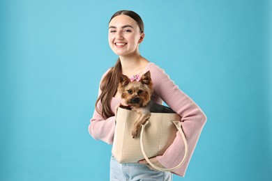 Beautiful young woman holding bag with cute Yorkshire Terrier dog on light blue background