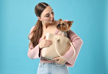 Beautiful young woman holding bag with cute Yorkshire Terrier dog on light blue background