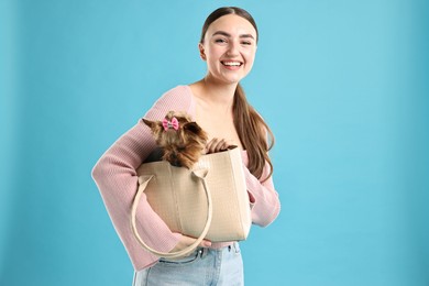 Beautiful young woman holding bag with cute Yorkshire Terrier dog on light blue background