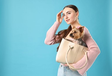 Beautiful young woman carrying cute Yorkshire Terrier dog in bag on light blue background, space for text