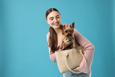 Beautiful young woman holding bag with cute Yorkshire Terrier dog on light blue background, space for text