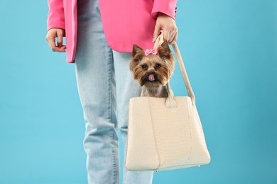 Woman carrying cute Yorkshire Terrier dog in bag on light blue background, closeup