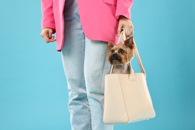 Woman carrying cute Yorkshire Terrier dog in bag on light blue background, closeup