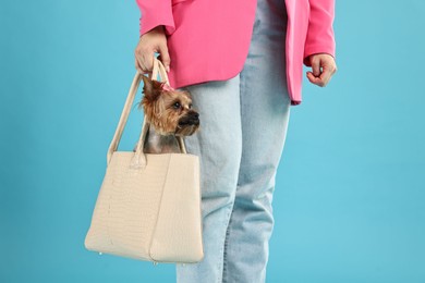 Woman carrying cute Yorkshire Terrier dog in bag on light blue background, closeup