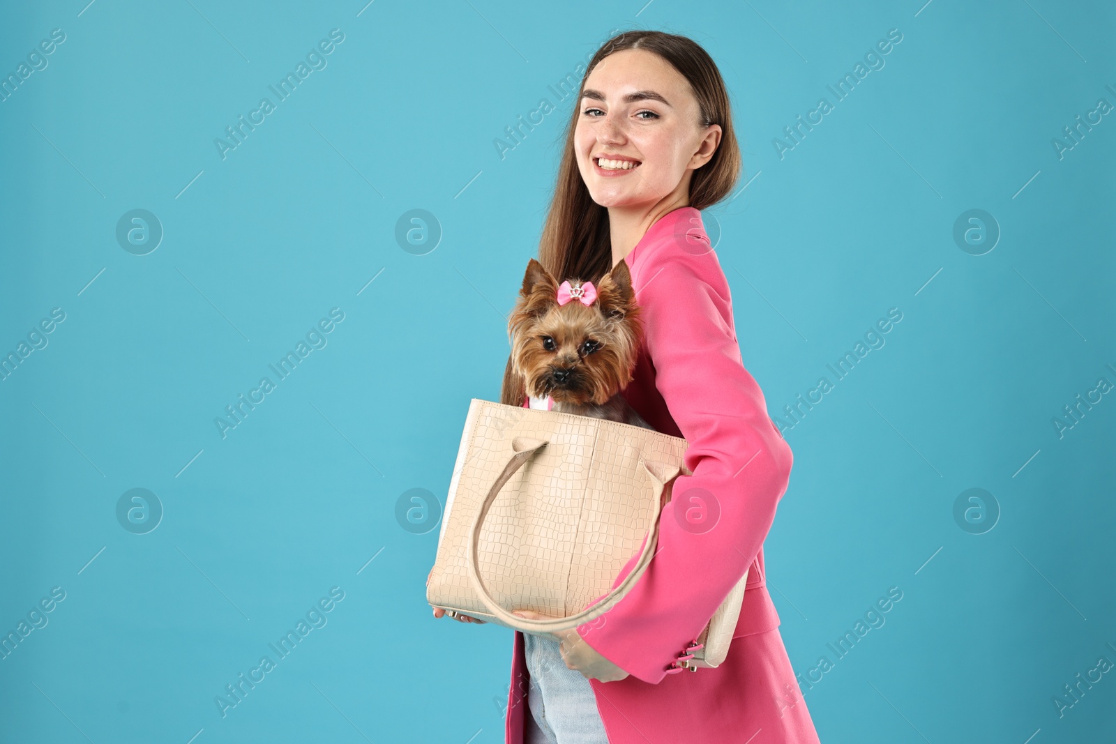 Photo of Beautiful young woman carrying cute Yorkshire Terrier dog in bag on light blue background