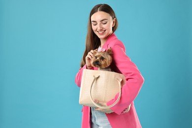 Beautiful young woman carrying cute Yorkshire Terrier dog in bag on light blue background