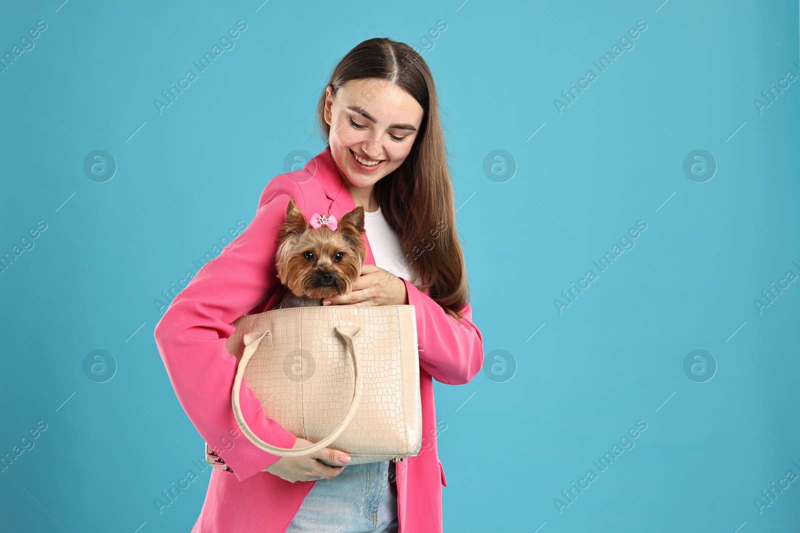 Photo of Beautiful young woman carrying cute Yorkshire Terrier dog in bag on light blue background, space for text